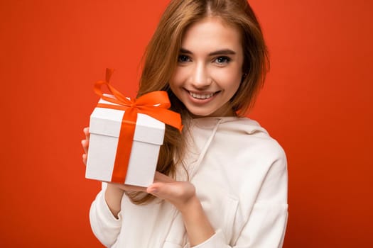 Portrait photo shot of pretty smiling cute funny blond young woman isolated over orange background wall wearing white hoodie holding stylish white gift box with red ribbon and looking at camera.