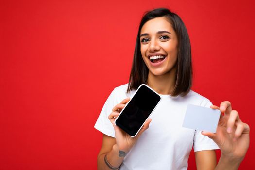Pretty happy woman wearing everyday stylish clothes isolated on background wall holding and using phone and credit card making payment looking at camera.