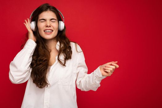 Attractive emotional young brunette female person wearing white shirt and optical glasses isolated over red background wearing white wireless bluetooth earphones listening to music with sincere emotions. free space