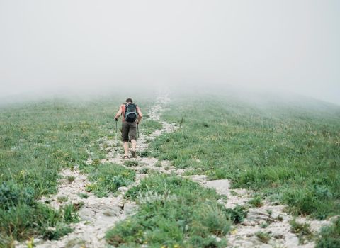 Hiker young man with backpack and trekking poles climbing up mountain in mist in summer outdoor, rear view