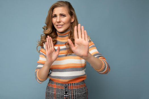 Portrait of young pretty beautiful sad angry dissatisfied blonde woman with sincere emotions wearing casual striped sweater isolated over blue background with copy space and showing no gesture and stop sign with palms. Negative concept.