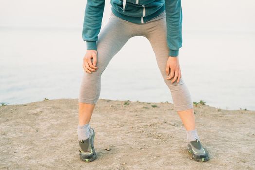 Fitness young woman warming-up her knee and preparing to run on coast in summer