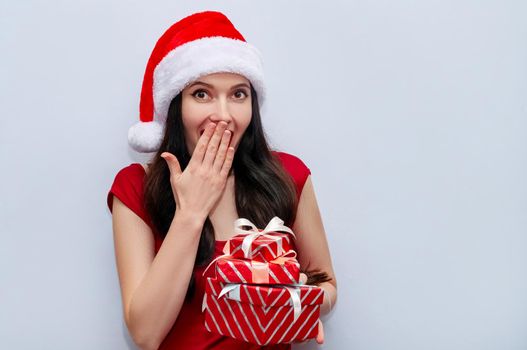 Close up Christmas Photo of Impressed Girl Screaming Wow OMG with Gift Boxes in Red Dress and Santa Hat on Grey Background.