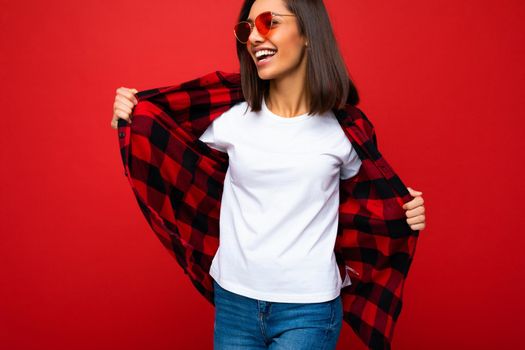 Photo of young beautiful happy smiling brunette woman wearing trendy white t-shirt with empty space for mock up and red check shirt and red sunglasses. Sexy carefree female person posing isolated near red wall in studio with free space. Positive model with natural makeup.