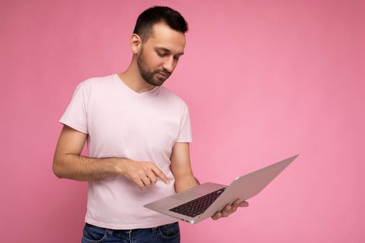 Handsome brunet man holding laptop computer and typing on the keyboard looking at netbook monitor in t-shirt on isolated pink background.