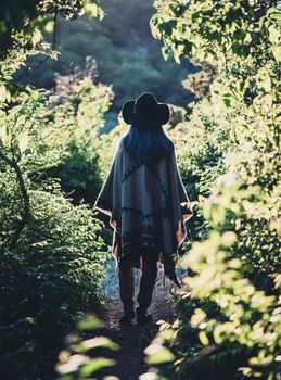 Fashionable young woman wearing in hat and poncho walking on path in summer park, rear view