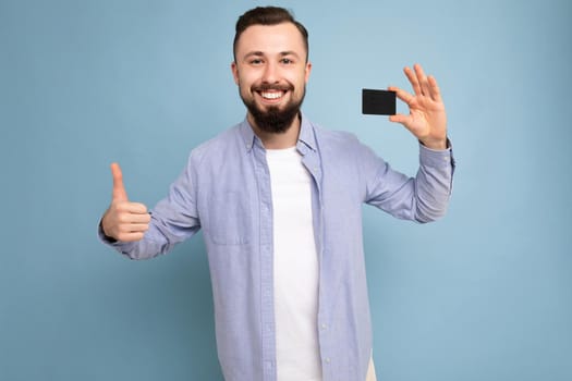 photo shot of Handsome smiling brunette bearded young man wearing stylish blue shirt and white t-shirt isolated over blue background wall holding credit card looking at camera and showing thumbs up gesture.