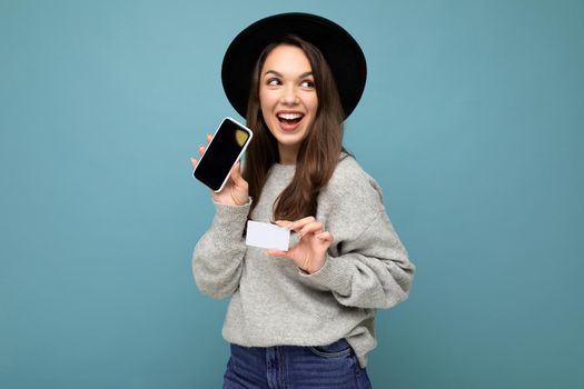 Photo of attractive joyful young brunette woman wearing black hat and grey sweater isolated over blue background holding credit card and mobile phone with empty display for mockup looking to the side.