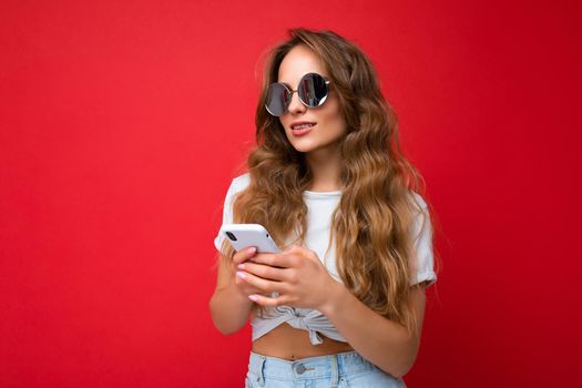Photo shot of attractive positive good looking young woman wearing casual stylish outfit poising isolated on background with empty space holding in hand and using mobile phone messaging sms looking to the side and thinking about something.