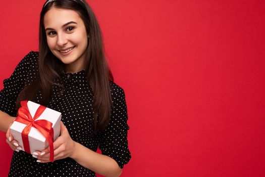 Closeup photo shot of beautiful happy smiling brunette female teenager isolated over red background wall wearing black blouse holding white gift box with red ribbon and looking at camera. Copy space