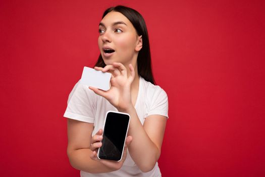 Photo of attractive happy young brunette woman wearing casual white t-shirt isolated over red background with empty space holding in hand mobile phone and showing smartphone with empty screen for mockup and credit card looking to the side.