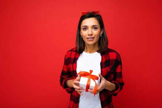 Photo shot of pretty positive young brunette woman isolated over red background wall wearing white casual t-shirt and red and black shirt holding white gift box with red ribbon and looking at camera.
