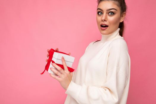 Closeup shot of attractive positive amazed young brunette woman isolated over pink background wall wearing white sweater holding gift box and unboxing present looking at camera.