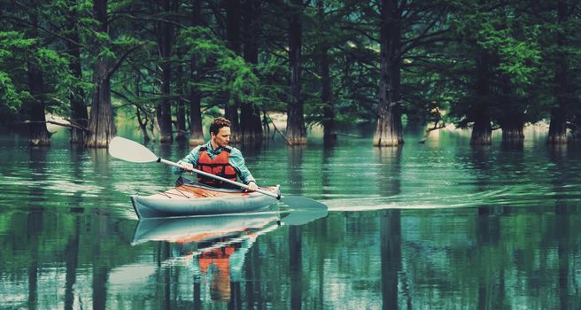 Young man paddling in kayak on lake on background of beautiful trees in summer. Toned image