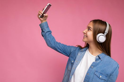 Side-profile Photo of beautiful positive smiling young dark blonde woman wearing blue jean stylish shirt and casual white t-shirt isolated over pink background wearing white bluetooth wireless earphones and listening to good music and using mobile phone taking selfie photo looking at gadjet.