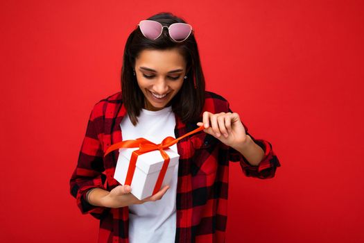Charming positive amazed young brunet woman isolated on red background wall wearing white casual t-shirt and stylish red and black shirt holding white gift box with red ribbon and unboxing surprise.