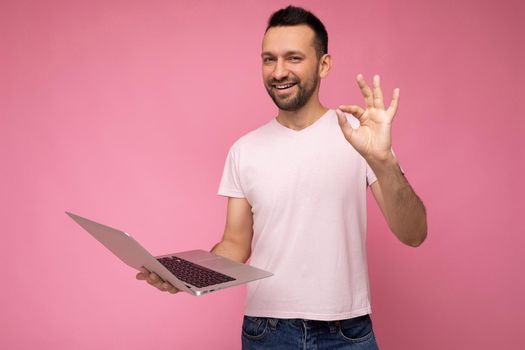 Handsome happy and funny brunet man holding laptop computer and showing gesture okay looking at camera in t-shirt on isolated pink background.