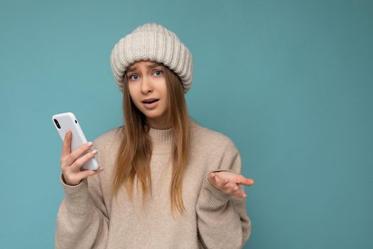 Photo of beautiful asking upset young blonde woman wearing stylish beige warm sweater and knitted winter warm beige hat isolated on blue background with empty space holding in hand and using mobile phone messaging sms looking at camera and having questions.