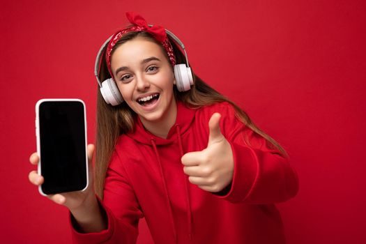 Attractive joyful smiling brunette girl wearing red hoodie isolated on red background holding and showing smartphone with empty display for cutout wearing white wireless headphones listening to funny music looking at camera and showing thumbs up gesture.