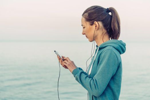 Fitness young woman in headphones chooses the music on her smartphone for workout on background of sea