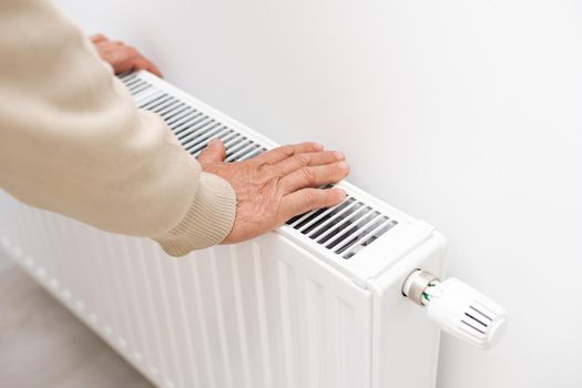 An elderly man froze in his hands, a man warms his hands near a heating radiator in an apartment, a man freezes in an apartment