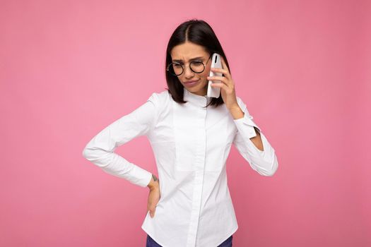 Beautiful dissatisfied young brunette woman wearing white blouse and optical glasses standing isolated over pink background talking on mobile phone looking down.
