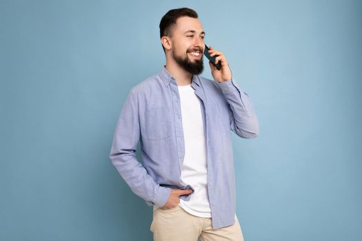 Side-profile Photo shot of good looking young brunet bearded man wearing casual blue shirt and white t-shirt poising isolated on blue background with empty space holding in hand and communicating talking on mobile phone looking to the side.