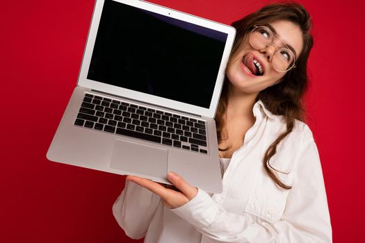 Crazy mad beautiful brunette curly young woman holding computer laptop wearing glasses white shirt showing tongue looking up to the side isolated over red wall background. Cutout
