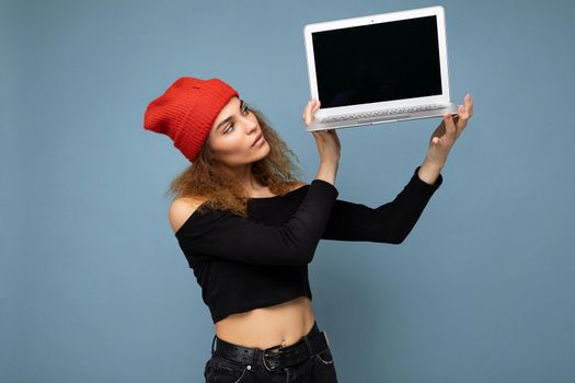 Close-up portrait of beautiful self-confident dark blond curly young woman holding laptop computer looking at netbook keyboard and monitor wearing black crop top and red and orange do-rag isolated over light blue wall background. Mock up, copy space, empty space
