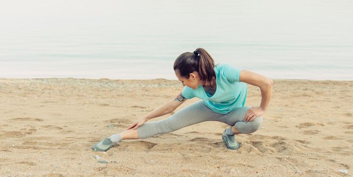 Fitness young woman stretching her legs and preparing to run on sand beach in summer, workout