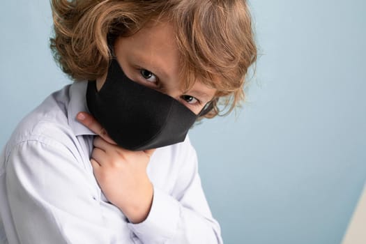 Closeup photo of male child in reusable virus protective mask on face against coronavirus isolated on the blue background wall.