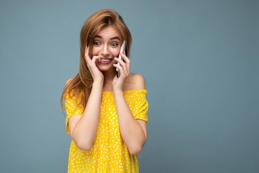 Closeup photo of pretty positive young blonde woman wearing stylish yellow summer dress standing isolated over blue background holding and talking on mobile phone with sincere emotions looking to the side.
