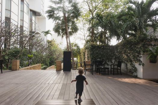 Back view of little boy running on wooden deck of hotel veranda with green palms around.