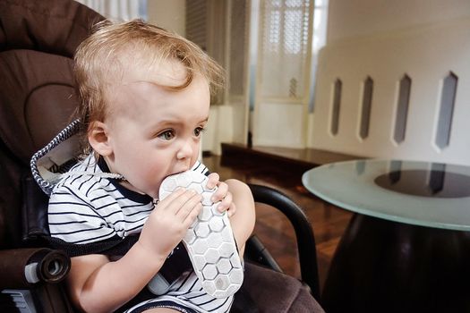 Little toddler sitting in chair and biting tiny shoe looking away.