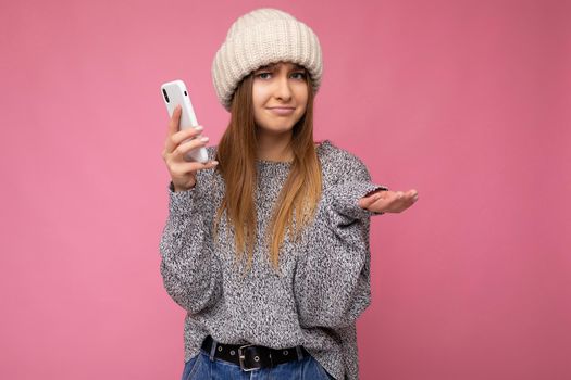 Beautiful funny asking young blonde woman wearing casual grey sweater and beige hat isolated over pink background holding in hand and using mobile phone looking at camera and having doubts.