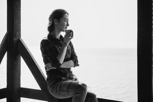 Smiling young woman sitting with cup in wooden arbor and enjoying view of sea. Monochrome image.