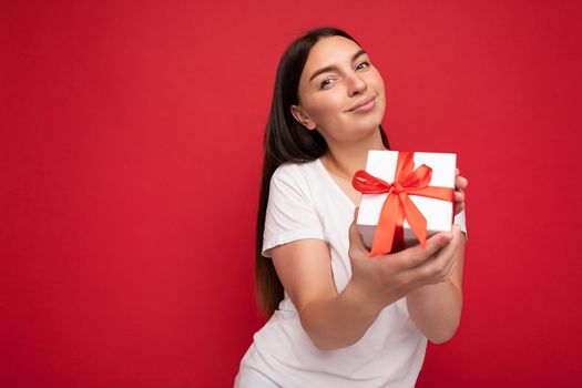 Photo of beautiful positive young brunette woman isolated over red background wall wearing white casual t-shirt for mockup holding white gift box with red ribbon and looking at camera. Free space