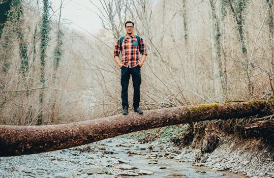 Hiker young man standing on fallen tree trunk over the mountain river in forest