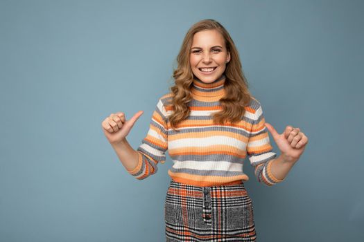 Young charming fascinating sexy positive happy smiling blonde woman wearing casual striped pullover isolated over blue background with free space and points fingers at herself.