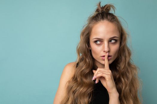 Photo of young beautiful blonde curly woman with sincere emotions wearing black top isolated over blue background with copy space and asking remain silent. Shhh gesture concept.