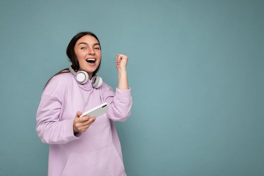 Beautiful happy smiling young brunette woman wearing purple hoodie isolated on blue background holding and using mobile phone surfing on the internet wearing white bluetooth headphones and having fun looking at camera and dancing. Copy space