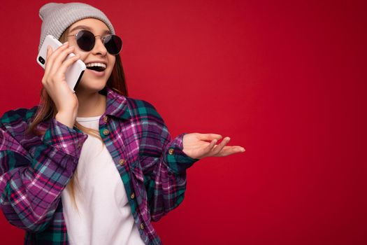 Closeup photo of beautiful happy positive young blonde woman wearing hipster purple shirt and casual white t-shirt grey hat and sunglasses isolated over red background holding in hand and communicating on mobile phone looking to the side and explaining something.