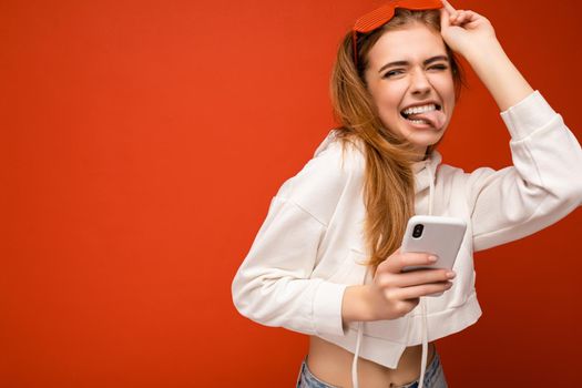 Beautiful emotional young blonde woman wearing stylish white hoodie and funny colorful glasses standing isolated over colourful background holding and using mobile phone communicating online on the internet and showing tongue. Copy space