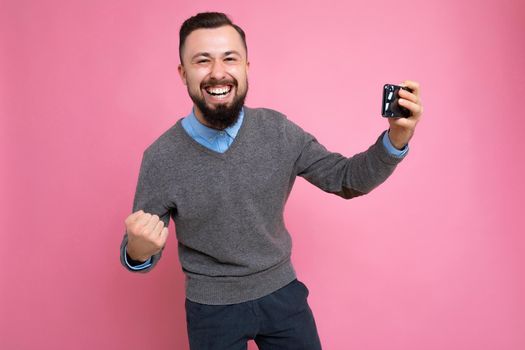 Overjoyed happy handsome young brunette unshaven man with beard wearing everyday grey sweater and blue shirt isolated on background wall holding smartphone watching videos via mobile phone looking at camera and celebrating winning.