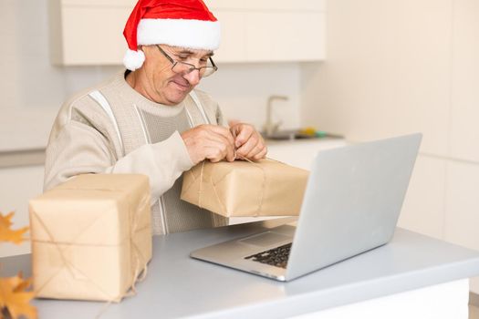 Senior man grandfather in Santa festive hat sitting on sofa and communicating with family online during videocall on laptop at Christmas decorated home. Pensioner congratulating children online