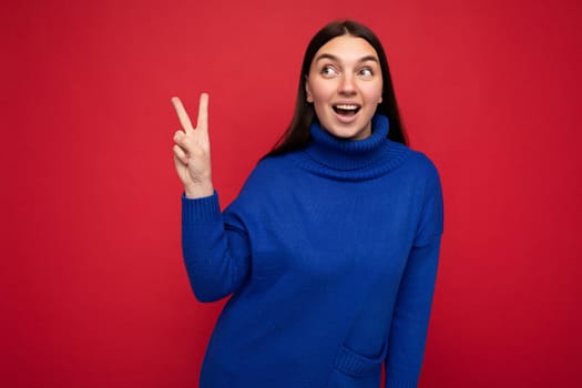 Photo of young positive happy smiling beautiful woman with sincere emotions wearing stylish clothes isolated over background with copy space and showing peace gesture with two fingers.