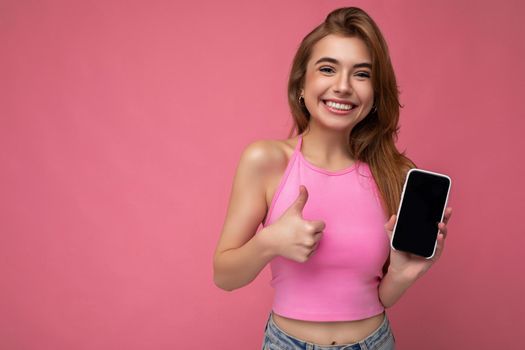 Beautiful joyful young blonde woman wearing pink top poising isolated on pink background with empty space holding in hand and showing mobile phone with empty display for mockup looking at camera and showing thumbs up gesture. copy space