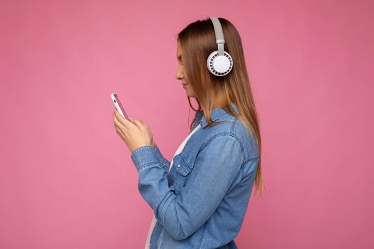 Side-ptofile closeup photo of beautiful young woman wearing stylish casual outfit isolated over colorful background wall wearing white wireless headphones and listening to music and using mobile phone messaging sms looking at smartphone display. copy space