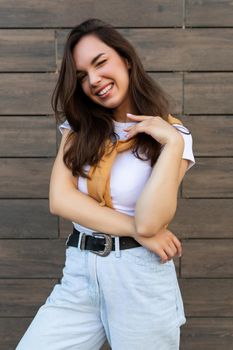 Vertical ptoto of young beautiful positive happy brunette woman standing against brown wall in the street and wearing stylish outfit.