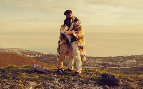 Travelers loving couple wrapped in plaid standing on peak of mountain above sea bay at sunset. Man embracing a woman